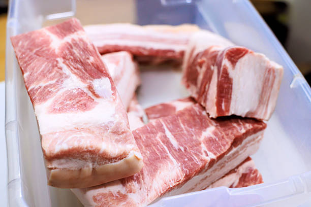 Several pieces of raw pork belly are arranged in clear plastic container on kitchen counter for cooking.