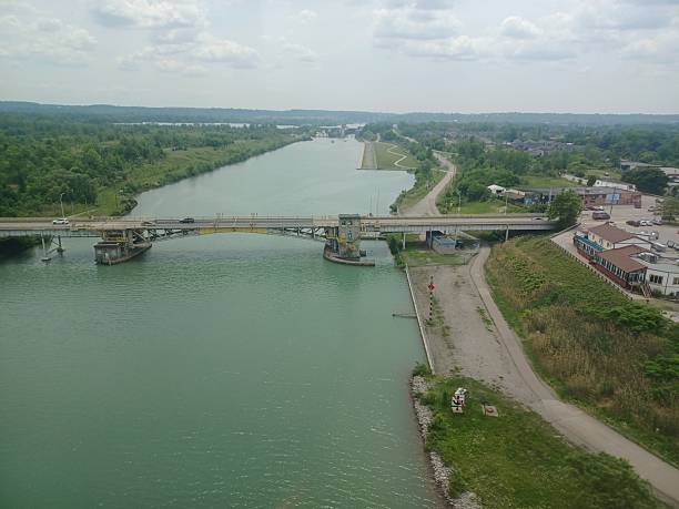 A road bridge spans a wide, greenish-blue water channel with surrounding green banks under a cloudy sky.
