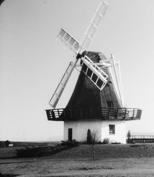A black and white photograph of a traditional windmill set in an open landscape, evoking a sense of history, craftsmanship, and rural heritage. The timeless structure reflects earlier methods of harnessing natural power, offering a quiet tribute to simpler days and enduring design.