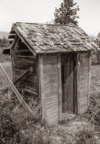 A black and white photograph of an old wooden outhouse standing as a quiet reminder of earlier rural life. The weathered structure reflects a time when daily routines were simpler, and amenities were modest, evoking nostalgia and the enduring character of days long past.