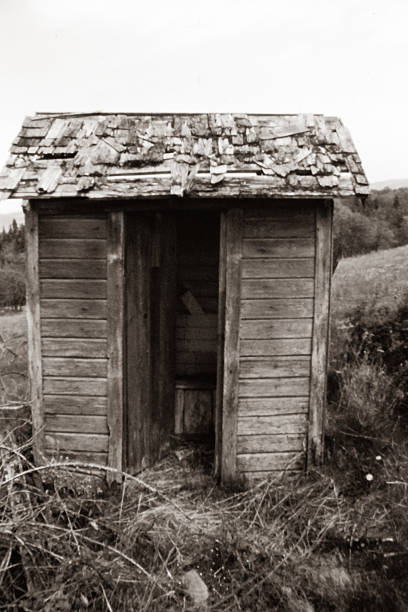 A black and white photograph of an old wooden outhouse standing as a quiet reminder of earlier rural life. The weathered structure reflects a time when daily routines were simpler, and amenities were modest, evoking nostalgia and the enduring character of days long past.