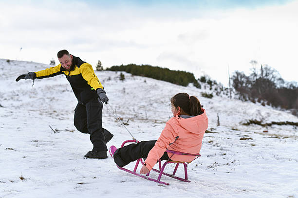 A man in a yellow and black jacket pulls a pink sled with a smiling young girl across a snowy hillside. The winter scenery and colorful outfits create a joyful family winter moment.