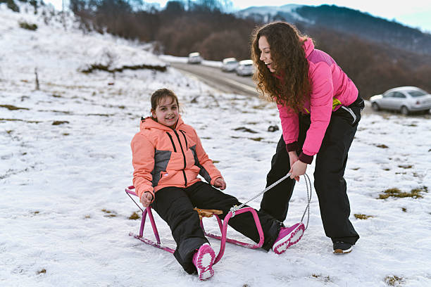 A young girl in a peach jacket and pink boots enjoys a playful sled ride on a snowy hillside, while her sister is helping, capturing a warm family moment in winter.