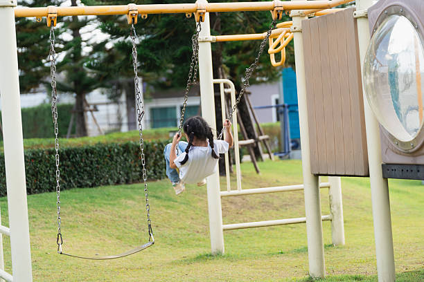 Young child playing joyfully on a swing in a vibrant playground surrounded by green grass and trees on a sunny day, promoting outdoor fun and happiness