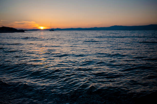 A serene sunset paints the sky with warm hues over the calm Adriatic Sea, with a distant sailboat adding to the picturesque scene