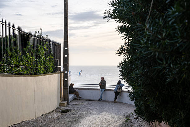 Estoril Lisbon District Portugal December 17 2025. People stand and sit at a coastal viewpoint overlooking the Atlantic Ocean during a winter afternoon.