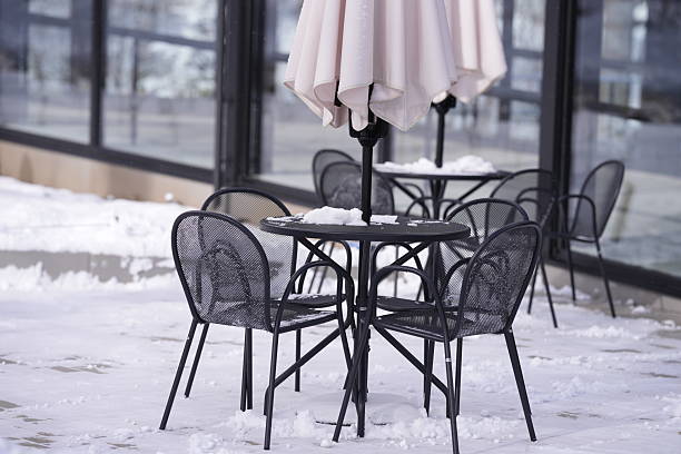 A table with four chairs and an umbrella is covered in snow. The chairs are black and the umbrella is white. The scene is set outside in the snow, with the chairs