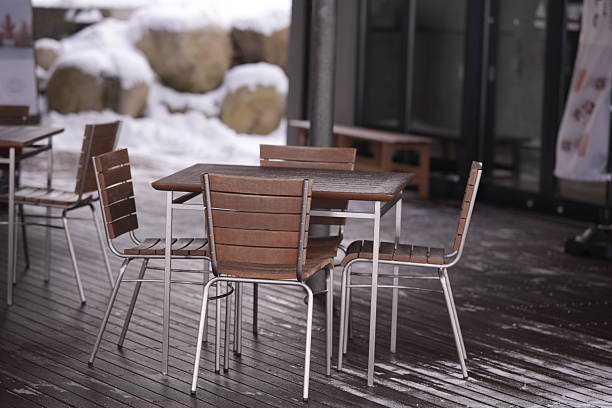 A wooden table with four chairs around it is empty. The chairs are made of wood and metal. The table is in a snowy area