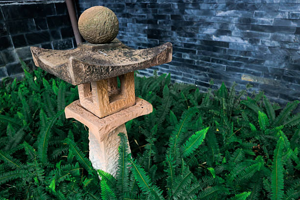 A shot of a traditional Japanese stone lantern (tōrō) nestled within a lush bed of vibrant green ferns. The background features a dark, textured slate stone wall.