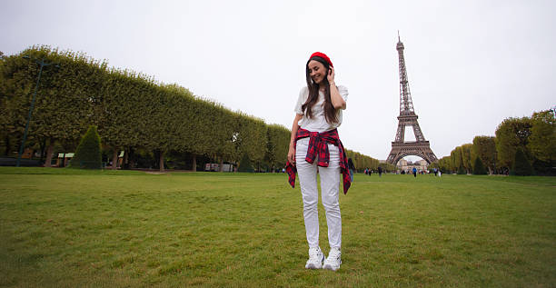 Young woman standing on green grass in a Paris park with the Eiffel Tower in the distance under a cloudy sky.