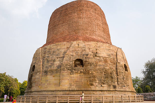 Sarnath, India - March 19, 2024: The Dhamekh Stupa, a large cylindrical brick and stone monument with carved stone details at the base, is shown with people walking nearby and trees in the background during daylight.