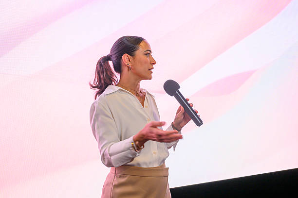 Woman on stage holding a microphone, delivering a confident, inspiring talk at a corporate conference or seminar, engaging an audience with ideas on leadership and innovation