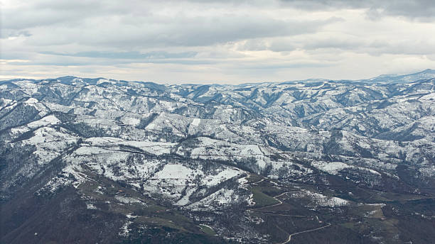 Aerial view of a vast mountainous landscape dusted with snow, revealing intricate patterns of hills, valleys, and winding roads under a dramatic cloudy sky