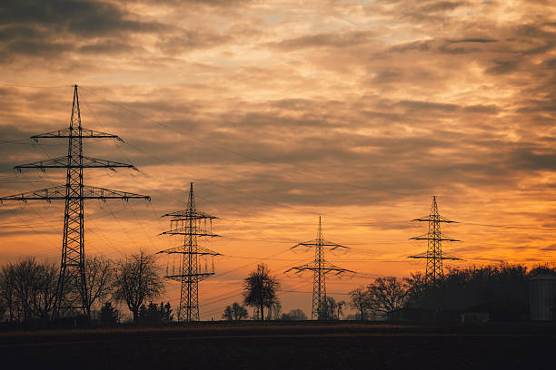 Electricity transmission towers and power lines silhouetted against a dramatic orange sunset sky. Energy infrastructure, electricity supply, and industrial landscape concept.