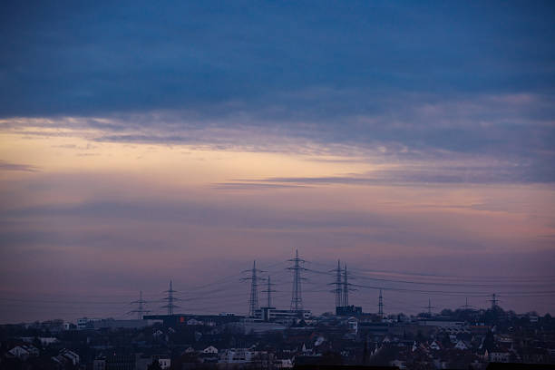 High voltage power lines stretching above an urban area during twilight, illustrating energy distribution, infrastructure, and the connection between city and industry.