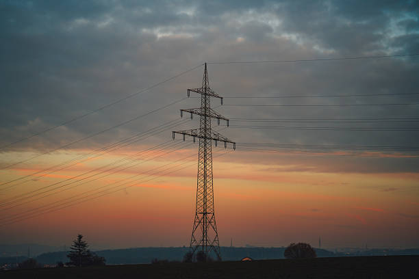Electricity transmission tower with overhead power lines silhouetted against a colorful dusk sky. Industrial energy infrastructure in a rural landscape, representing power distribution, energy supply, and modern utilities.