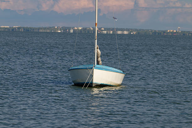 A solitary sailboat rests on the tranquil surface of a large body of water, with a blurred cityscape visible on the horizon under a cloudy sky