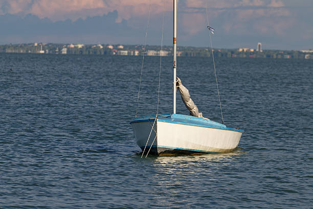 A solitary sailboat rests on the tranquil water, its mast reaching towards a cloudy sky, with a blurred urban landscape visible on the horizon