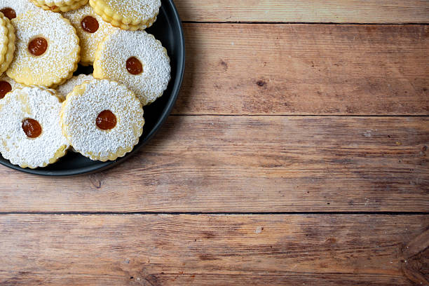 Top view of traditional Linzer cookies dusted with powdered sugar on a black plate over rustic wooden table. Clean copy space for text and seasonal food design.