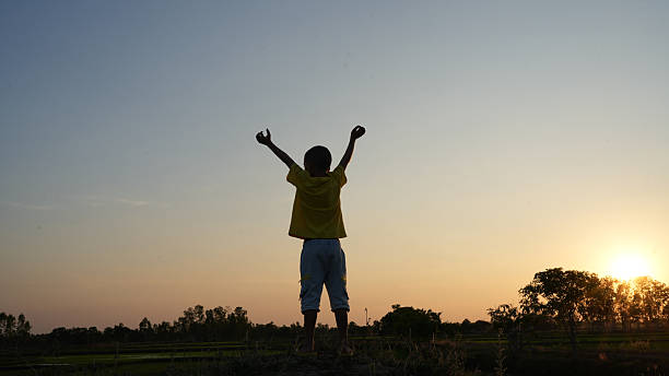 Child stands with arms raised in celebration against sunset backdrop, embodying joy and freedom in serene landscape