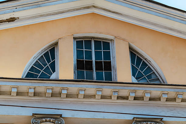 Closeup shows arched attic window with divided panes under triangular pediment. Decorative cornice and trim details frame roofline.