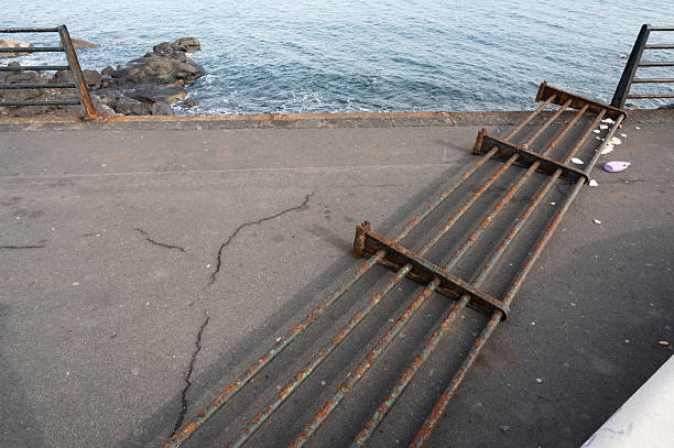 A rusted metal railing lies collapsed on a seaside promenade in Catania following Cyclone Harry. Cracks are visible in the pavement, with scattered debris including a purple plastic item and white fragments. The walkway overlooks a rocky shoreline and the ocean, highlighting structural damage and safety hazards caused by the storm.