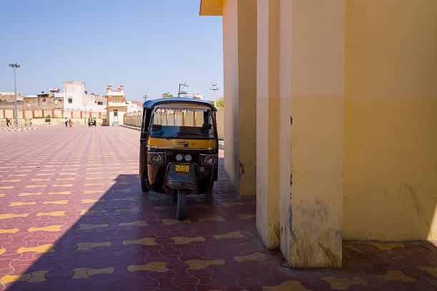 Pushkar, India - March 17, 2022: An auto rickshaw is parked next to a yellow building in a large, mostly empty paved parking area with another rickshaw and a few people visible in the background.