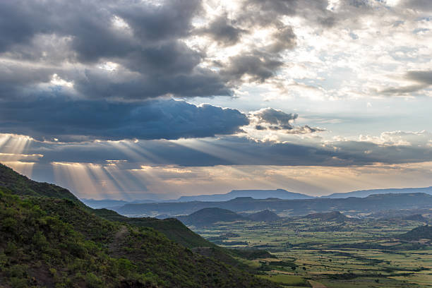 A wide landscape shot captures the breathtaking beauty of the Ethiopian Highlands near sunset. Golden sunbeams, or crepuscular rays, pierce through a heavy layer of dark storm clouds, illuminating the rolling green hills and patchwork agricultural fields below. The scene conveys a powerful sense of majesty and hope, showcasing the vast, rugged topography and dramatic weather patterns typical of the Horn of Africa's high-altitude plateaus.