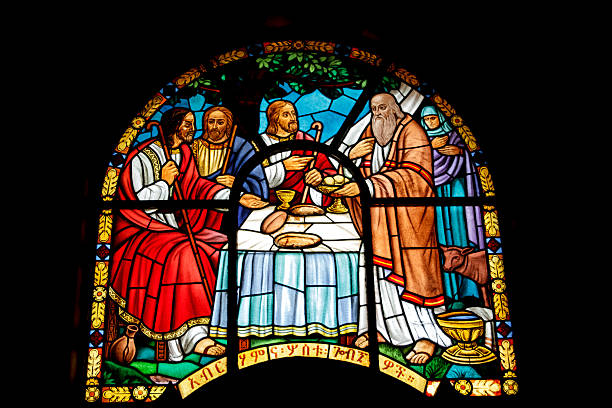A vibrant, high-contrast photograph of a stained-glass window within the Holy Trinity Cathedral. The scene illustrates the biblical story of Abraham hosting three divine visitors at a table set with bread and wine. Rich primary colors and traditional Ethiopian Orthodox iconography define the piece, which includes Ge'ez script at the base. The image captures the artistic heritage and spiritual storytelling found in one of Ethiopia's most significant religious landmarks.