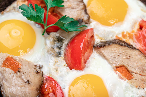 A close-up of a delicious, rustic breakfast featuring fried eggs cooked in a black cast iron skillet with slices of homemade roasted pork (Buzhenina) and fresh cherry tomatoes. The dish is garnished with parsley and served on a wooden board.