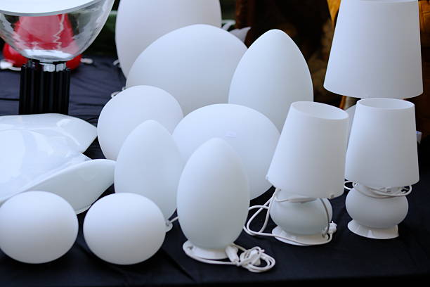 Assorted modern table lamps and light fixtures displayed on a table, featuring minimalist shapes and frosted white surfaces. The clean forms and soft tones convey contemporary design, interior decor, lighting concepts, and home accessories. No people visible.