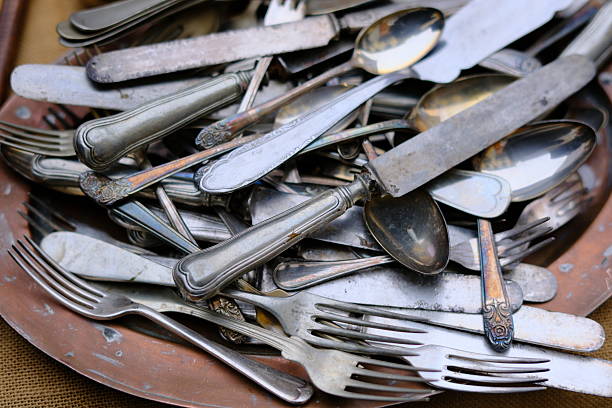 Assorted vintage knives, forks, and spoons stacked together in a metal tray. Worn surfaces and tarnished silverware create a rich texture that evokes history, reuse, craftsmanship, and second-hand household items. No people visible