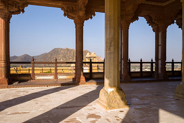 Amber, India - March 16, 2022: The view from the Diwan-e-aam pavilion at Fort d'Amber shows carved stone columns, a balustrade, yellow stone buildings, and arid hills under a clear blue sky.
