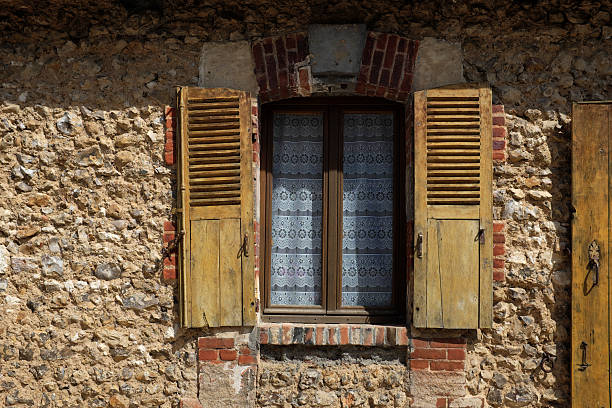 A rustic window with wooden shutters on a stone wall, featuring a lace curtain behind the glass