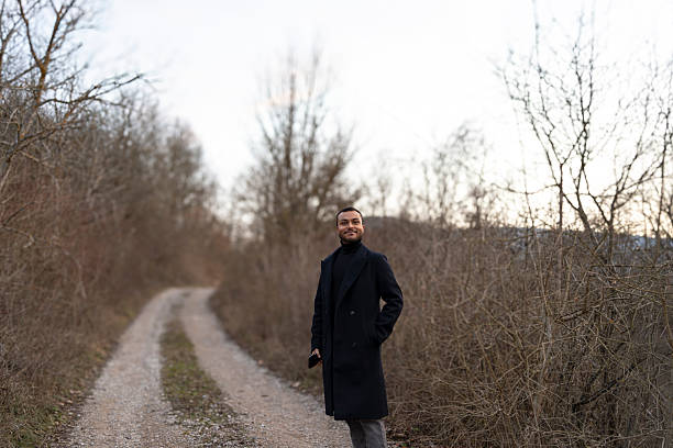 Young man smiling, holding smartphone, standing on dirt path in rural nature during winter