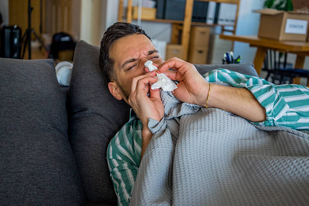 istock Young man lying on a sofa, feeling sick with a cold, blowing his nose and using nasal drops, covered with a blanket, showing signs of fever. 2258130371