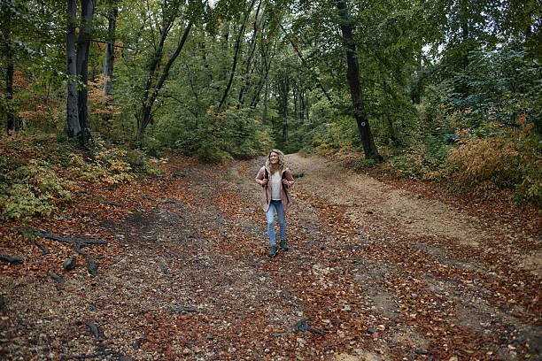 A winding footpath, partially covered with fallen autumn leaves, leads into a serene winter forest. On the right, a distinctive birch tree with white bark features a small orange trail marker, alongside a rustic wooden railing. The surrounding trees are bare, their intricate branches forming a canopy against an overcast sky, creating a quiet and inviting atmosphere for a walk in nature.