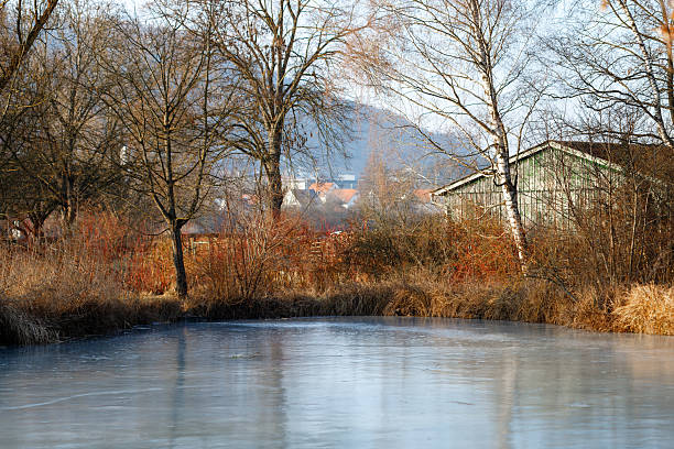 Winter scene showing frozen lake, bare trees, and an old barn near residential area