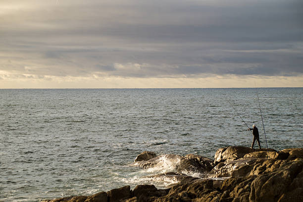 Wide shot of a man fishing on large sea rocks under a dramatic cloudy sky during sunset.
