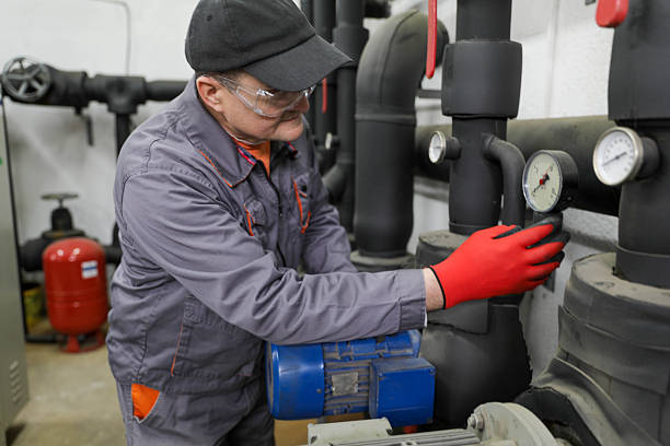 Mature Industrial Technician Wearing Protective Gloves And Safety Glasses Inspecting Electric Motor And Pressure Gauges In Mechanical Room, Performing Maintenance And Monitoring Industrial Equipment, Demonstrating Professional Technical Work, Safety Standards, And Industrial Infrastructure Management.