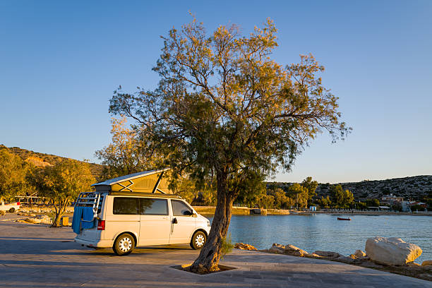 Port de Marathi, Greece - October 23, 2022: A white camper van with an elevated roof is parked beside a tamarisk tree along the paved waterfront at Port de Marathi, with calm water, distant hills, and coastal buildings visible in the background during golden hour.