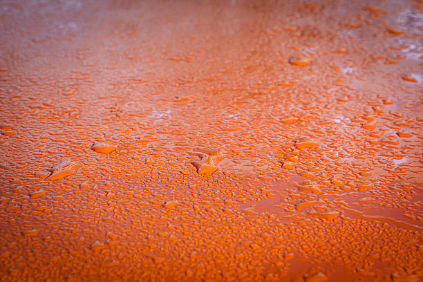 Macro image of numerous water droplets covering a bright orange surface, creating a rich texture and dynamic visual contrast. The vivid color and glossy appearance evoke freshness and highlight the interaction between liquid and solid material.