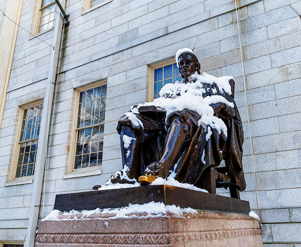 Cambridge, Massachusetts, USA - December 21, 2024: Close-up of the John Harvard statue covered in snow in Harvard University's Harvard Yard. The John Harvard monument (c. 1884) is a bronze statue of John Harvard (1607-1638) by Daniel Chester French. The monument inscription is \