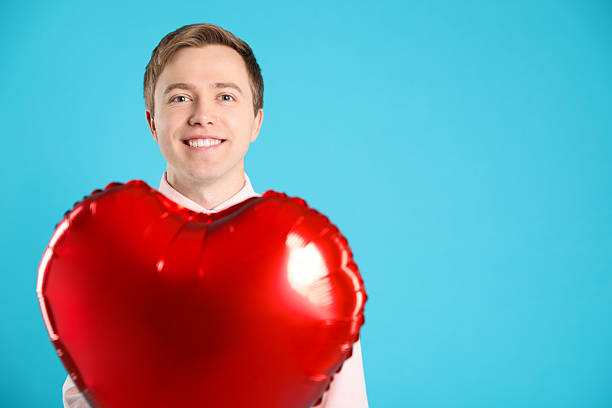 Cheerful man with heart shaped balloon on light blue background, space for text. Romantic gift