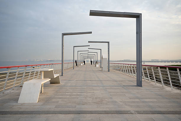 A long, modern seaside walkway features a series of repeating steel frames that create a striking geometric perspective leading toward the horizon. The clean lines and minimalist design of the structure contrast beautifully with the natural backdrop of calm water and distant city skyline. White benches and red railings add subtle color accents, while a few pedestrians stroll along the path, emphasizing scale and human interaction with public space. This image captures the harmony between urban architecture and coastal nature, ideal for illustrating themes such as sustainable urban design, contemporary public infrastructure, and architectural photography