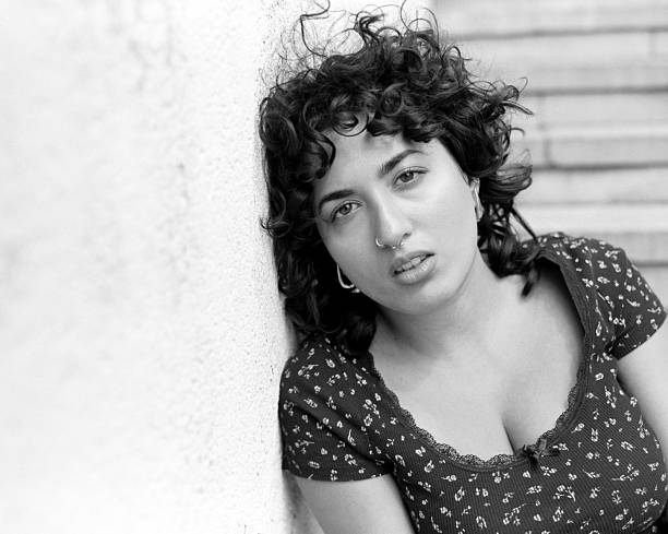 Young adult woman with curly dark hair and nose ring leaning against a wall, capturing a melancholic look with a vintage black and white film aesthetic, reflecting emotional depth and individuality