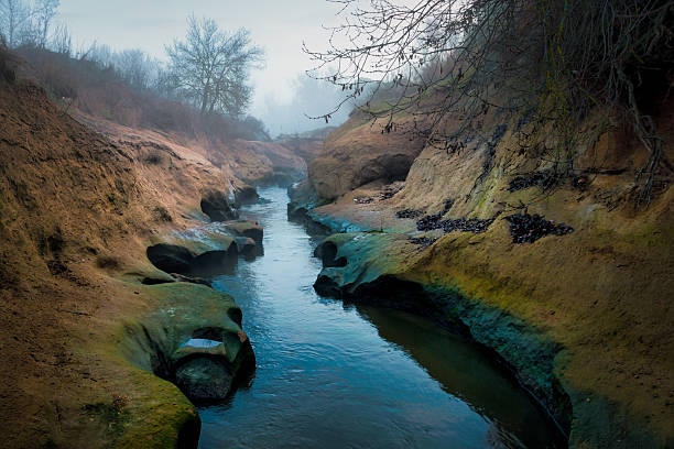 Narrow river channel flowing smoothly between eroded rock formations covered with moss. The calm water surface and sculpted stones create a serene, natural atmosphere that evokes time, erosion, continuity, and quiet strength. Ideal for concepts related to nature, mindfulness, resilience, natural processes, and environmental balance.