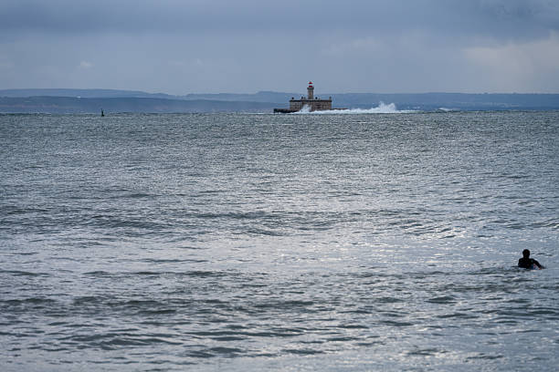 Oeiras Portugal November 21 2025. Waves break around an offshore lighthouse visible on the horizon during cloudy weather.