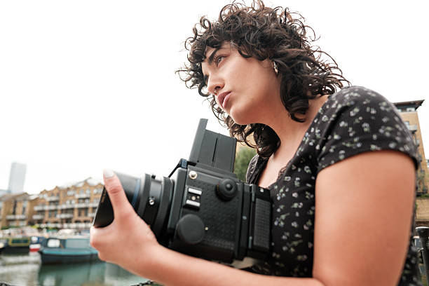 Young woman photographer composing a shot with a mamiya rb67 medium-format film camera outdoors by a city canal, focused on traditional analog film photography craft