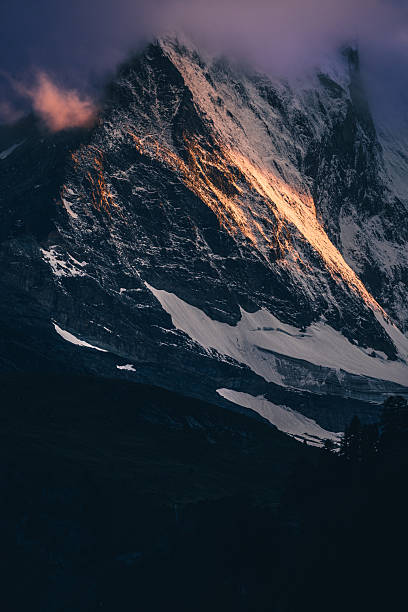 A dramatic close-up view of the Matterhorn in Zermatt, captured under low clouds with patches of snow clinging to its steep, angular rock face. Warm alpenglow light grazes the mountain’s surface, creating a strong contrast against the dark, shadowed slopes below.